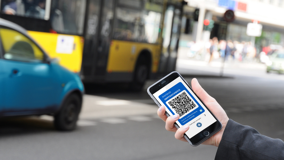 Eine Hand vor einem Bus hält ein Smartphone mit einem Impfzertifikat auf dem Screen.