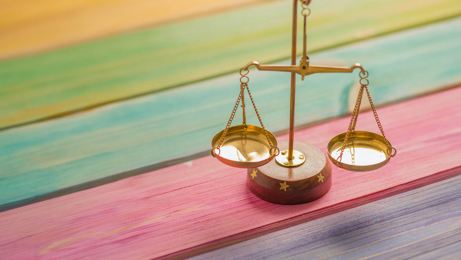 Eine kleine goldene Waage auf einem Holztisch dessen Holzleisten regenbogenfarben angemalt sind und für Chancengerechtigkeit steht