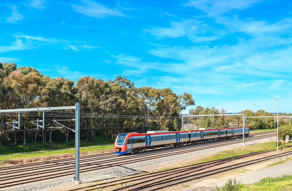 Ein rot-silberner Keolis Zug fährt vor Bäumen und einem strahlend blauen Himmel von rechts nach links.
