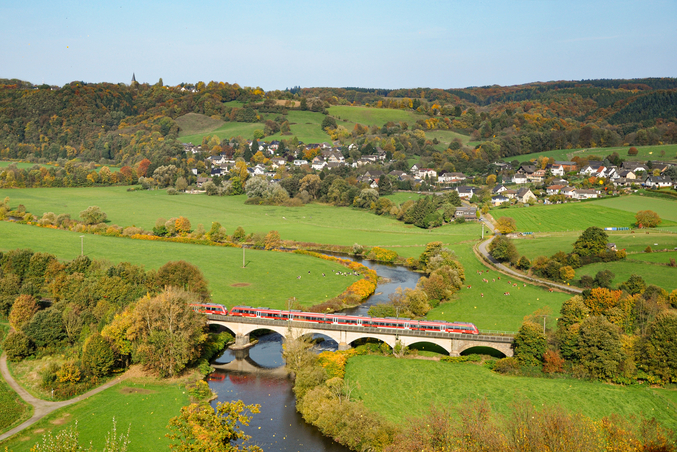 Ein roter DB Regio Zug fährt über eine Brücke über einem Fluss im Grünen mit einer Kleinstadt im Hintergrund.
