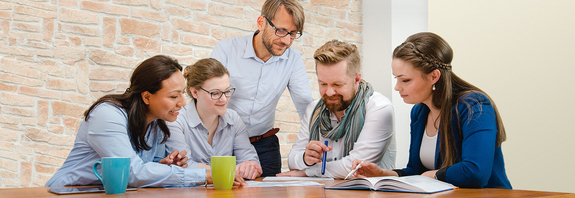 Fünf Kollegen sitzen an einem Tisch und diskutieren die darauf liegenden Dokumente