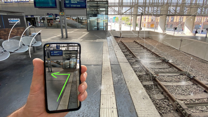 Auf einem Bahngleis hält eine Hand hält ein Smartphone, das einen Weg auf dem Bahngleis per Pfeil anzeigt.