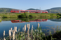 Ein roter DB-Regionalzug fährt an einem Fluss vorbei und wird in diesem gespiegelt.