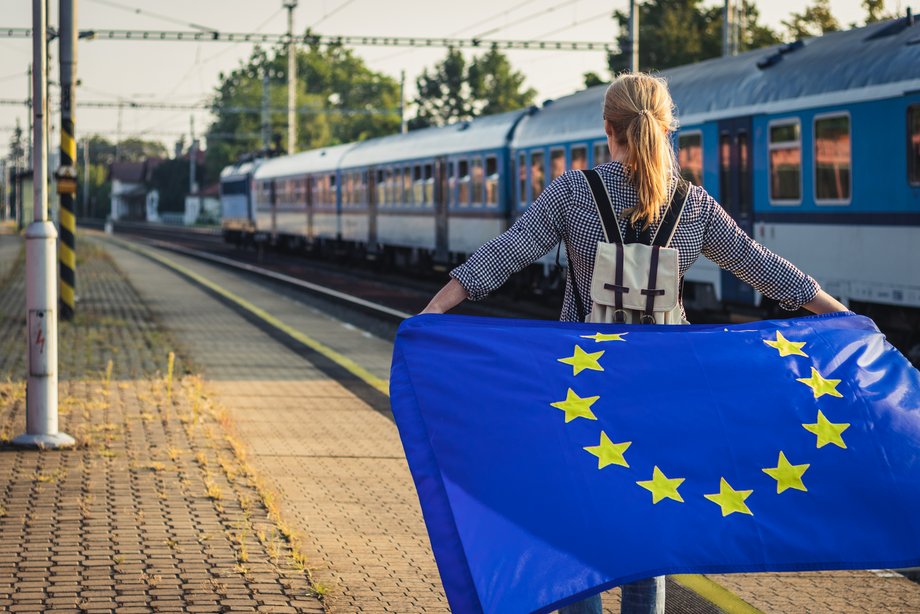 Frau mit Europa-Fahne am Bahnhof, vorbeifahrender Zug – Symbol für europäische Trassenbestellung nach TEL TSI